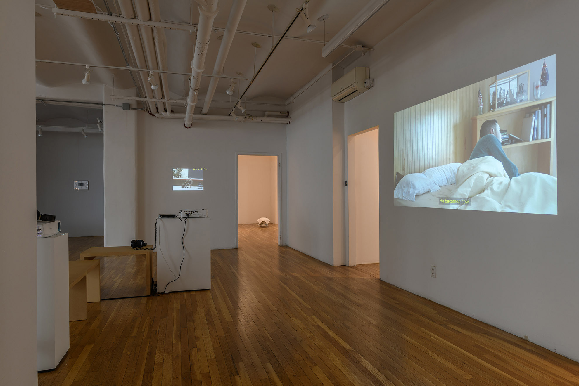 [A view of the main gallery showing in greater detail one of the color video projections of a person in bed. Receding in space are two illuminated open doorways, another color projection nearby, and two white plinths supporting projects on either side of two benches.]